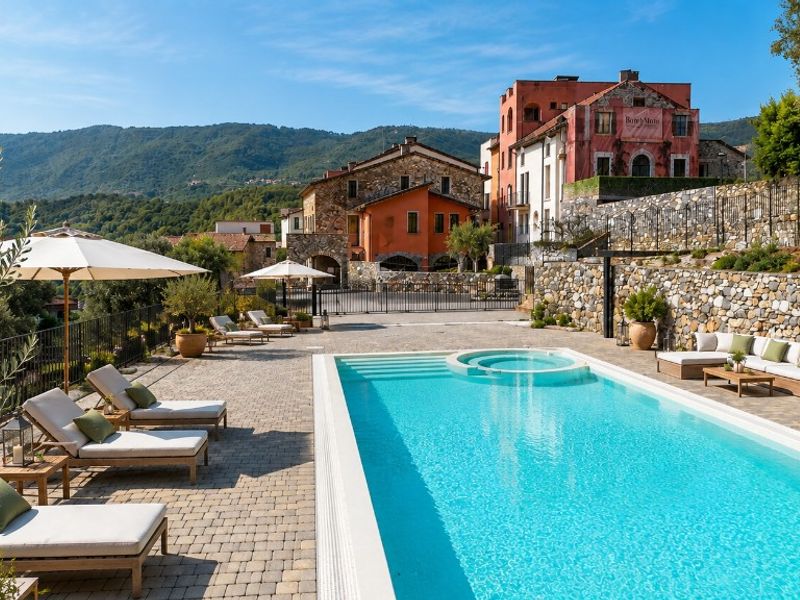 Borgo Agosti ad Andora — Area piscina con lettini, divano da esterno e vista sulle colline e gli edifici in pietra