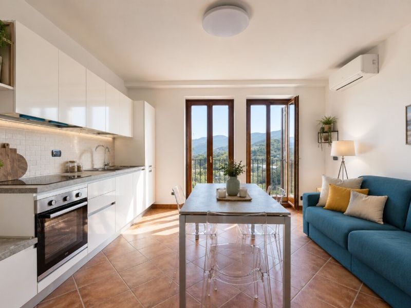 I Borghi a Boissano — Cucina moderna bianca con open space, divano azzurro e porte-finestre con vista sulle colline